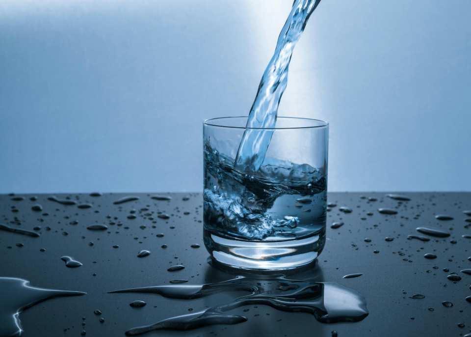 Clear glass of fresh, clean water being poured, with droplets splashing on a dark surface, symbolizing the pure, refreshing water delivered by whole-house water filters in Massachusetts homes